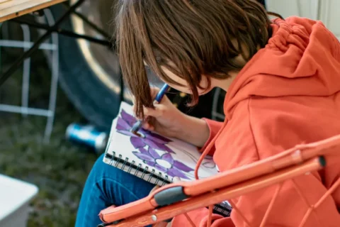 A child with short brown hair, wearing a bright orange hoodie, sits in an orange folding chair while focused on painting. They are using a paintbrush to add purple colour to a floral drawing in a spiral-bound sketchbook. The scene is outdoors, with a bicycle wheel visible in the blurred background.