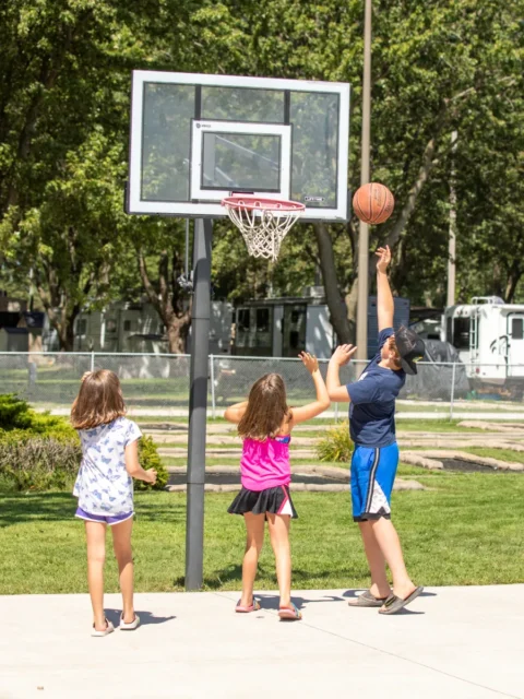 Three children play basketball on an outdoor concrete court on a sunny day. A boy in a blue shirt and shorts reaches up to shoot a basketball toward a clear backboard and hoop, while two young girls watch from the court. The background features a grassy area with trees and several parked RVs behind a chain-link fence.