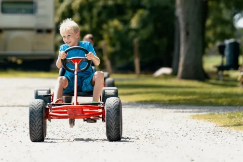 A young blond boy in a blue t-shirt smiles while driving a red pedal go-kart on a gravel path. He has both hands on the steering wheel. The scene is set outdoors with green trees and a camper in the soft-focus background.
