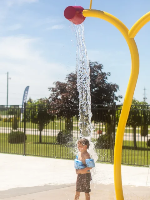 A young boy wearing blue swim floaties and dark trunks stands on a concrete splash pad as a stream of water pours over his head from a red bucket. The bucket is part of a yellow, curved water-play structure. In the background, a black metal fence separates the splash pad from a grassy area with trees under a bright, sunny sky.