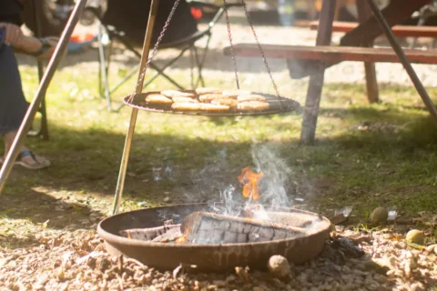 Several hamburger patties cook on a circular metal grate suspended by chains from a tripod over an open wood fire. The fire burns in a shallow metal pit on the ground, with smoke and orange flames rising toward the meat. The background shows a blurred campsite with a picnic table and lawn chairs on a sunny day.