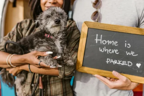 A close-up shot of a couple standing in front of a vehicle, with the woman holding a small, scruffy grey dog. The man holds a small wooden-framed chalkboard that has the phrase "Home is where we PARKED" written in white chalk with a small heart. Only the couple's torsos and hands are visible, emphasizing the dog and the sentiment on the sign.