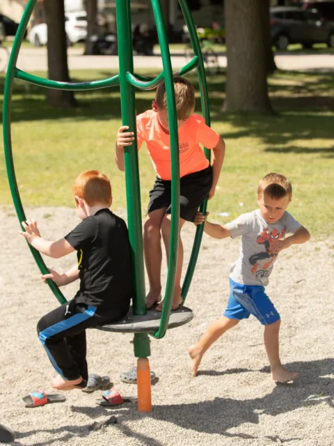 Three young boys play on a green, spinning playground structure over a pea gravel surface. One boy in a black t-shirt sits on the base, another in a bright neon-orange shirt stands on the platform holding the centre pole, and a third boy in a grey Spiderman shirt runs alongside to push the spinner. Several pairs of sandals are scattered on the ground nearby.