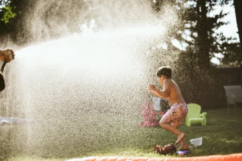 A young boy in pink-patterned swim trunks jumps and dances through a bright, misty spray of water from a garden hose on a sunny day. The water droplets catch the sunlight, creating a glowing effect across the centre of the image. The scene is set in a green backyard with trees and a small lime-green lawn chair in the background.
