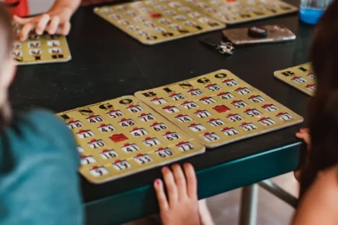 A close-up, slightly high-angle shot of people playing Bingo at a dark table. Several golden-tan Bingo cards with sliding red shutters are spread across the surface. In the foreground, a child's hands rest on the edge of the table near two cards, while other players' hands and cards are visible in the soft-focus background.