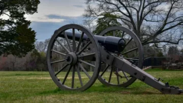 A large, historic black iron cannon rests on a carriage with two tall wooden wheels in a grassy field. The cannon is positioned on a slight incline, pointing toward the left of the frame. In the background, bare trees stand against a cloudy, late-afternoon sky, giving the scene a quiet and sombre atmosphere.