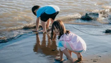 Two children play on a sandy shoreline as gentle waves wash in. A young girl in a pink shirt and colourful leggings crouches in the foreground, looking toward the camera with a smile, while a boy in a blue shirt bends over in the background to touch the water. Their reflections are visible in the wet, dark sand under the bright daylight.