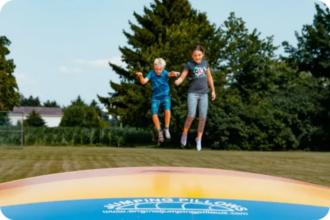 Two children jump high into the air on a large, yellow and orange inflatable jumping pillow. The activity is taking place outdoors on a sunny day with a green field and trees in the background.
